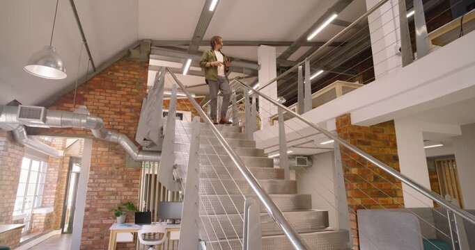 After checking tablet, African American adult man descending metal stairs in loft in olive jacket