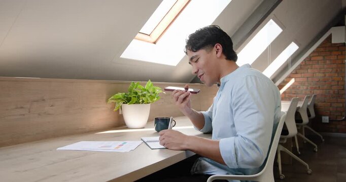 Adult man right at counter lifting smartphone, speaking while noting charts in notebook, copy space