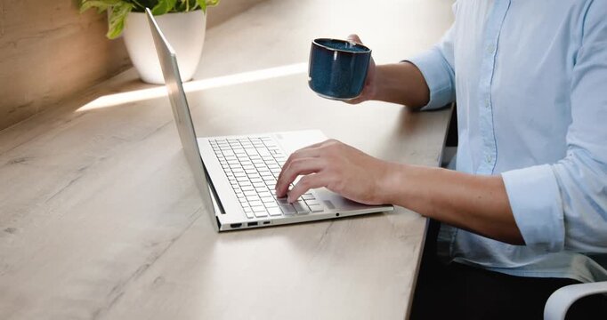 Asian man typing on laptop at desk, pausing, sipping from blue mug, resuming typing, copy space