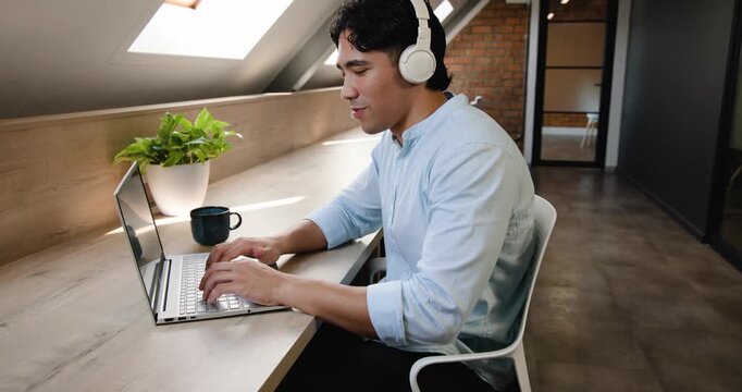 Man in blue shirt with white headset typing on laptop under skylights, sipping mug, finishing task