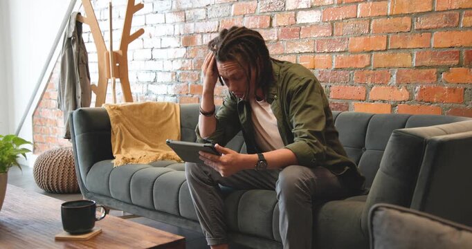 African American man sitting on green sofa glancing left, shifting to tablet and studying content