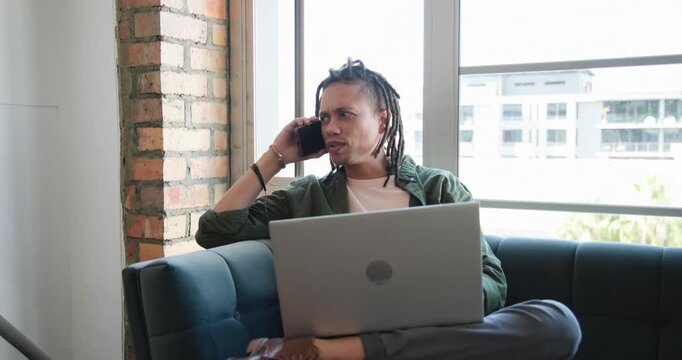 Adult African American man taking incoming call, laughing, typing on laptop, working on blue couch