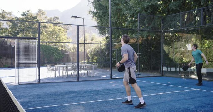 Diverse men returning high lob on blue court, shaking hands at net, walking with paddles