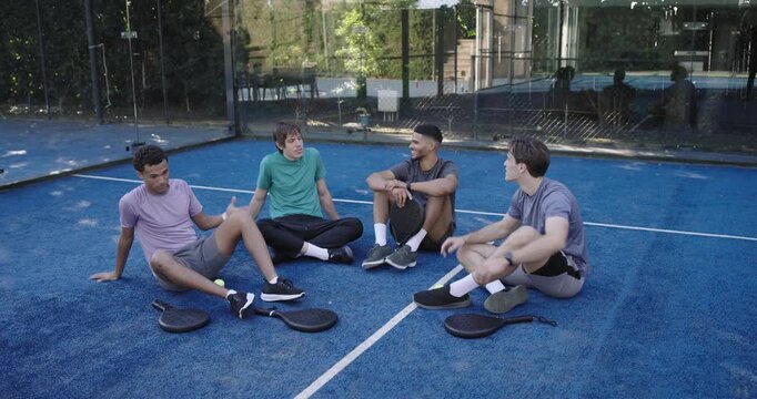 Diverse male friends sitting on court after playing, passing paddles and ball while debriefing game
