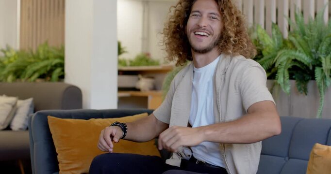Adult man responding to camera cue, smiling then laughing, leaning back on tufted sofa with plants
