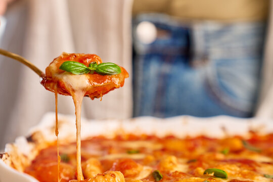 Gnocchi in tomato sauce served on spoon with cheese stretching from baking dish and basil on top