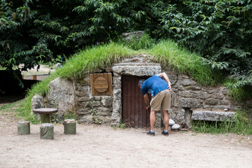 Fototapeta premium A male tourist bends down to explore the miniature wooden door of a whimsical stone earth house with a grass roof during a family summer vacation.
