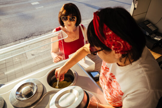 Curious mature woman watching female vendor scoop gelato at sunny outdoor ice cream kiosk