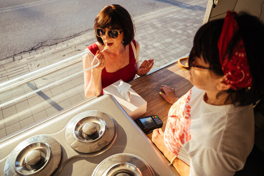 Mature woman tasting ice cream sample while speaking with vendor at sunny summer kiosk