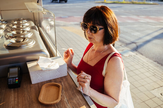 Satisfied mature woman evaluating gelato sample at sunny outdoor ice cream kiosk counter