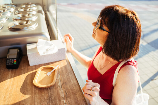 Curious mature woman checking gelato flavors before ordering at sunny street food kiosk