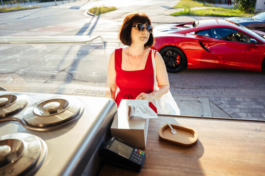 Stylish mature woman waiting for gelato beside red sports car on sunny summer city street