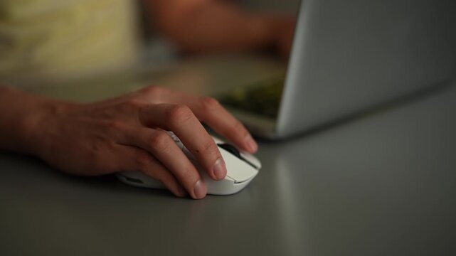 Close-up video of persons hand using white wireless mouse. User clicks and scrolls working on laptop in dark room. Screen light reflects on desk, creating dynamic mood. Shooting in slow motion.