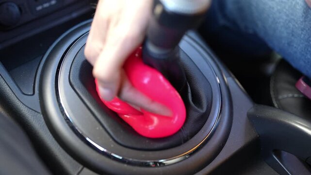 Top view of person hand pressing pink cleaning slime into crevices of car gear stick console, effectively collecting dust and debris from hard to reach areas for spotless interior.