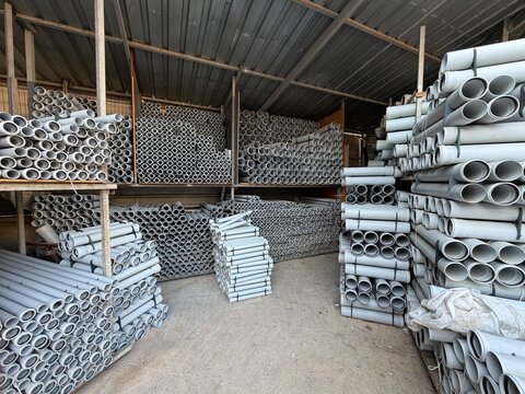 Warehouse storage scene with horizontal rows of light gray PVC pipes on wooden shelving, open ends facing camera, while upper stacks show slight outward tilt and gentle realignment