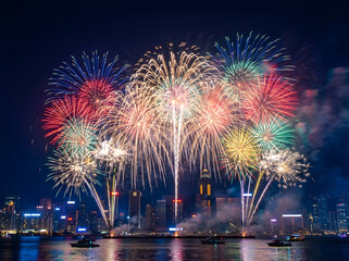 Fototapeta premium Colorful fireworks display over the victoria harbour skyline in hong kong at night with illuminated skyscrapers and boats on the dark water surface