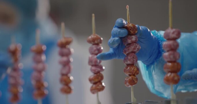 Raw sausage skewers standing upright in rows while a gloved hand picks one in a food production area