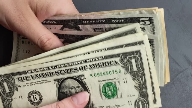Close-up of hands counting small denomination US dollar bills on a graphite grey background.