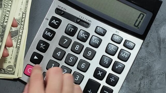 Close-up of hands counting small denomination US dollar bills and using a calculator on a graphite grey background.