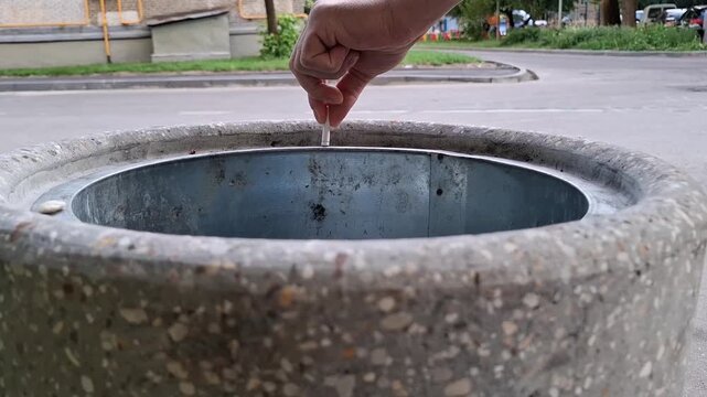 Man drops a cigarette butt into a concrete trash can on an outdoor city street. smoking habits, throwing garbage, city litter bin, urban pollution.