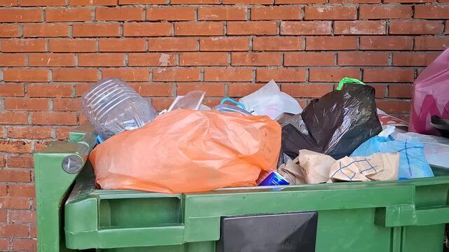 Green plastic dumpster is overflowing with plastic bags and bottles against a red brick wall outdoors. garbage sorting, waste management, municipal pollution, residential trash.
