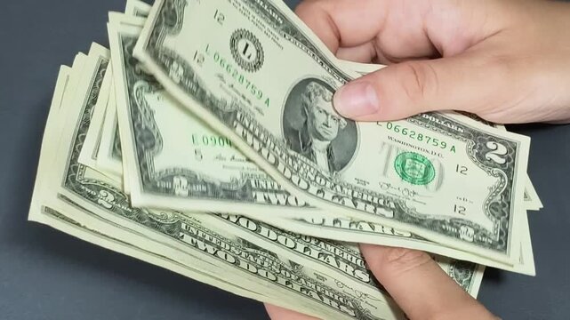 Close-up of hands counting two-dollar bills on a graphite grey background.
