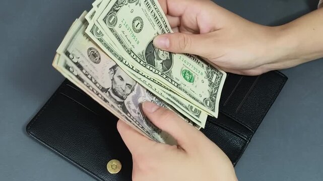 Close-up of hands counting US dollar bills and putting cash into a black wallet on a graphite grey background.