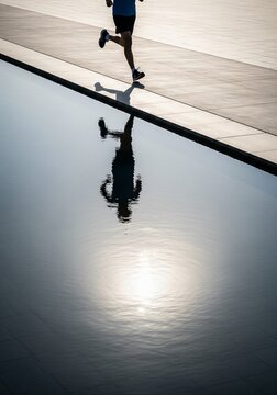 High angle view of a person running on a concrete path next to a water surface reflecting the runner and the bright sun.