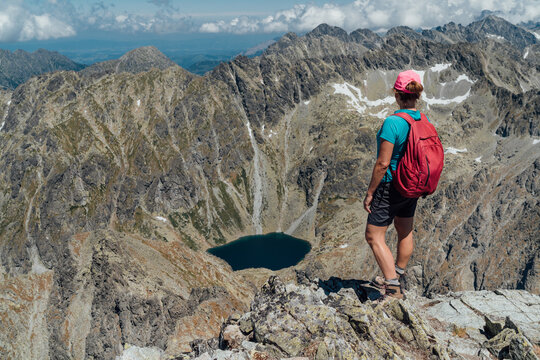 Woman trekker standing on rocky peak of Krivan mountain looking at mountain lake pleso, female hiker with backpack enjoying scenic view of High Tatras in Slovakia during summer hiking