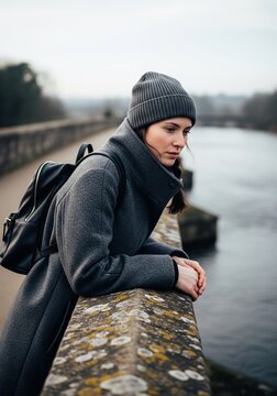 A young woman wearing a gray coat and a beanie hat leaning on a stone bridge and looking down thoughtfully