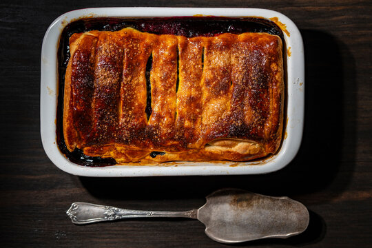 Homemade berry pie in baking dish with vintage spatula, closeup. Top view of freshly baked golden berry cobbler or puff pastry pie in white ceramic dish on dark wooden table with vintage cake server