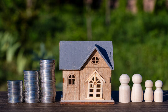 Wooden house toy placed between stacks of silver coins and symbolic wooden family of four on table, closeup. Real estate investment, mortgage planning, finance growth, and housing budget concept