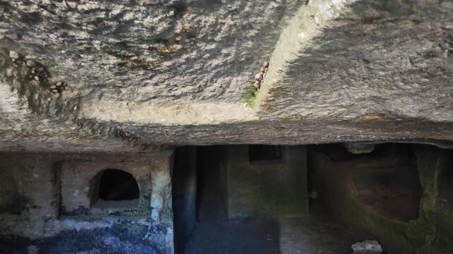 Salina, Malta - a panning shot across the rough, rock-cut ceiling of an ancient Paleo-Christian catacomb reveals arched openings and dark burial chambers below within the underground complex.