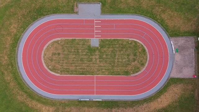 Top-down aerial drone footage of a small red athletics running track, showcasing the oval track layout, surrounding green sports field