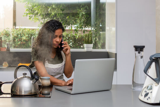 Serious woman using laptop while talking on smartphone in modern kitchen