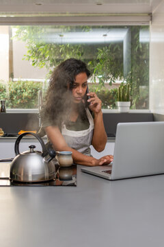 Concerned Hispanic woman talking on smartphone while using laptop in kitchen