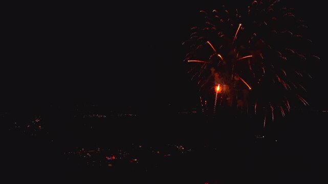 Bright fireworks light up the night sky over the city. People gather below to watch and enjoy the colorful bursts. The event takes place during a festive holiday celebration.