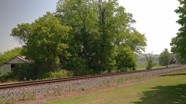 A steam train approaches along the tracks beside tall trees and a small building. The sun shines overhead in the afternoon sky. The area around is green with grass and bushes.
