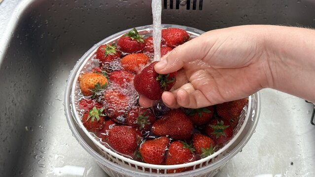 Hand rinses fresh strawberries in a colander under running tap water in a kitchen sink, clean food prep scene with bright red fruit, healthy home cooking mood