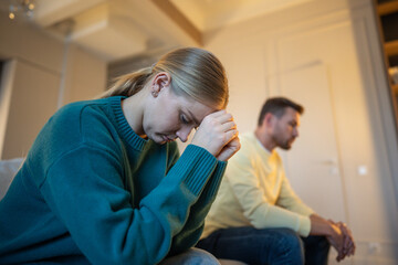 Fototapeta premium Heartbroken sad woman sitting on couch with distanced partner, relationships crisis and brawl, married couple after emotional quarrel, coldness and mistrust between husband and wife due to conflict.