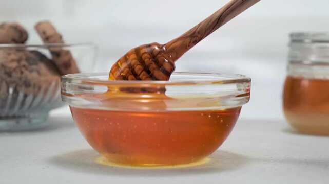 Golden honey flowing from wooden dipper into transparent glass bowl, slow motion