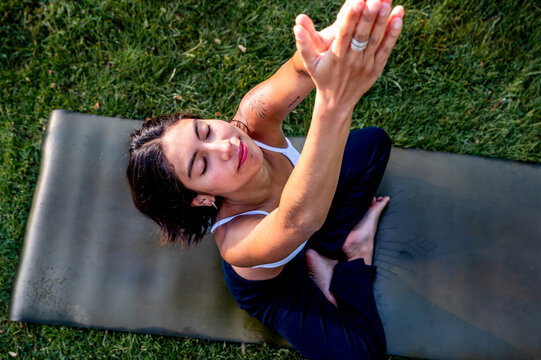 A young Hispanic woman practises a reclining stretch on a yoga mat in a lush green park at golden hour, a practice of mindfulness and mental wellbeing in the tranquil evening.