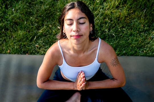 A young Hispanic woman practises seated meditation with prayer hands surrounded by greenery in a park at golden hour, a mindful summer fitness and wellbeing practice amid green summer nature.