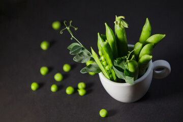 Fresh green pea pods in a white cup with scattered seeds on a dark background © Denis
