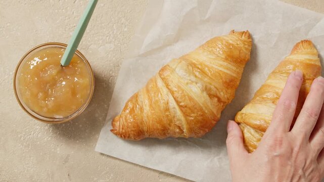 Top view of a freshly baked golden butter croissant and a small bowl of jam on baking paper placed on a light pastel beige kitchen table. A human hand gently places a second croissant beside them