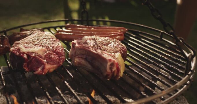 Seasoned steaks and sausages cooking on a round hanging grill grate over an open flame in a garden