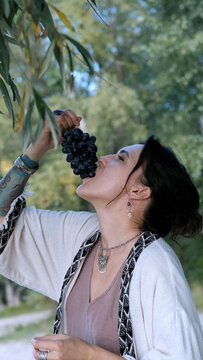 A beautiful brunette in a bohemian style relaxes outdoors in summer against a backdrop of a green forest, holding a large bunch of juicy black grapes