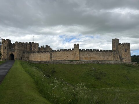 Historic stone castle wall and battlements viewed across a grassy green moat under a cloudy sky