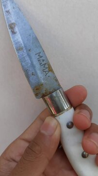 A hand holding an old, vintage folding pocket knife with a white handle and a stained metal blade against a neutral background.