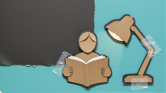Cardboard cutout of person reading book with desk lamp, taped to teal background.
